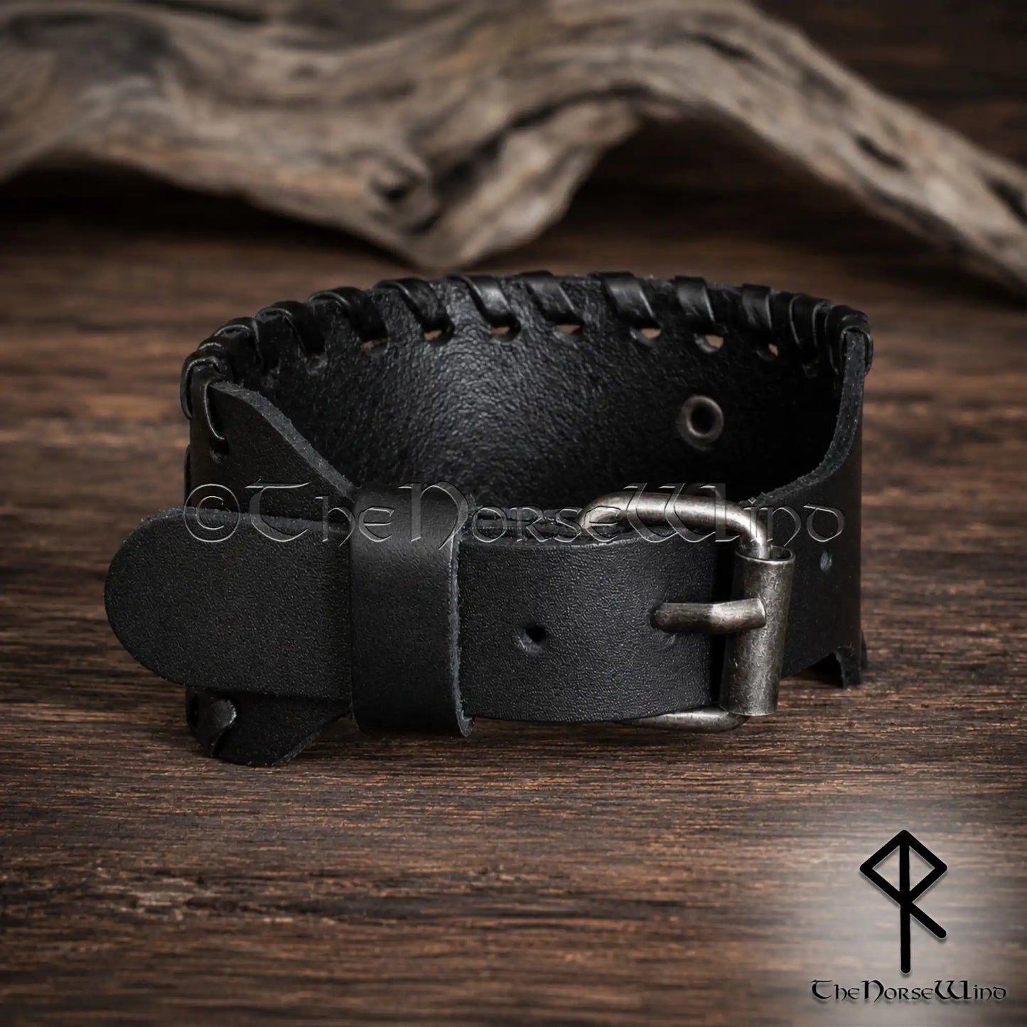 A black leather Viking-style cuff bracelet with a silver-toned metal buckle, displayed on a rustic wooden surface with a piece of driftwood in the background.