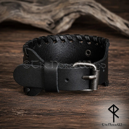 A black leather Viking-style cuff bracelet with a silver-toned metal buckle, displayed on a rustic wooden surface with a piece of driftwood in the background.