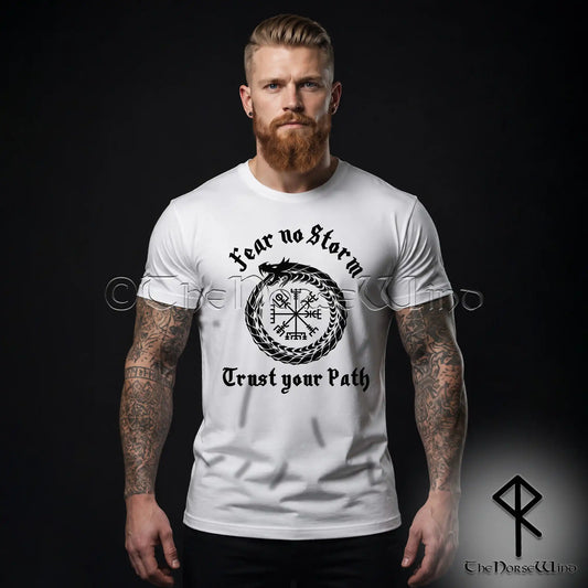 A man with a beard and tattoos wearing a white viking t-shirt with a black Norse design featuring the Midgard serpent, Vegvisir compass, and the text 'Fear no Storm' and 'Trust your Path'. The background is solid black.
